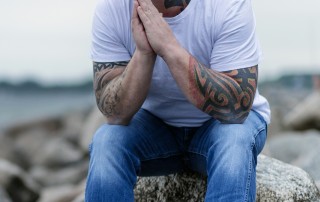 man with tattoos sitting on large rock thinking