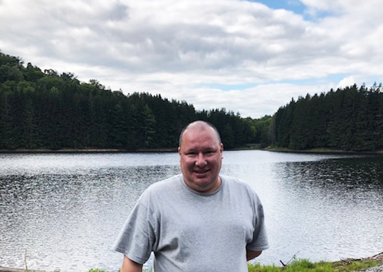 smiling man in grey shirt standing in front of lake - SJI Graduate Kris