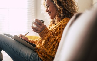 happy woman writing in journal and drinking coffee on sofa - new year