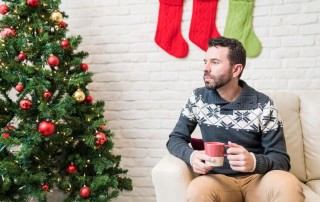 man in his thirties sitting alone next to Christmas tree drinking coffee or cocoa, thinking - loneliness