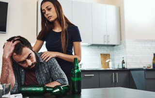 tired looking woman with her hands on the shoulders of her man who is sitting at a table full of empty beer bottles - codependent relationships
