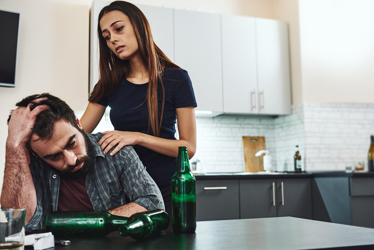 tired looking woman with her hands on the shoulders of her man who is sitting at a table full of empty beer bottles - codependent relationships