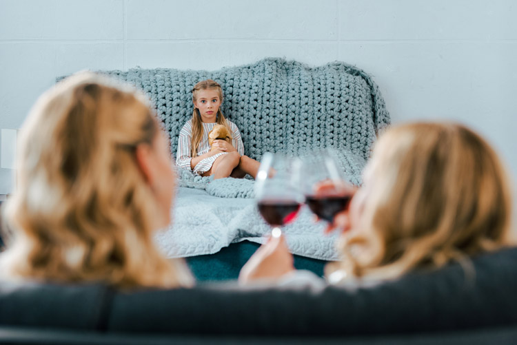 two women clinking wine glasses while a little girl looks on from the bed - wine mom culture