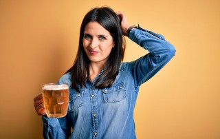 pretty brunette woman in a denim shirt holding a large mug of beer and scratching her head as she considers drinking it - gray area drinking