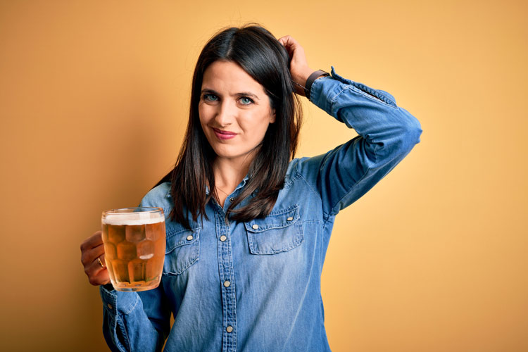 pretty brunette woman in a denim shirt holding a large mug of beer and scratching her head as she considers drinking it - gray area drinking