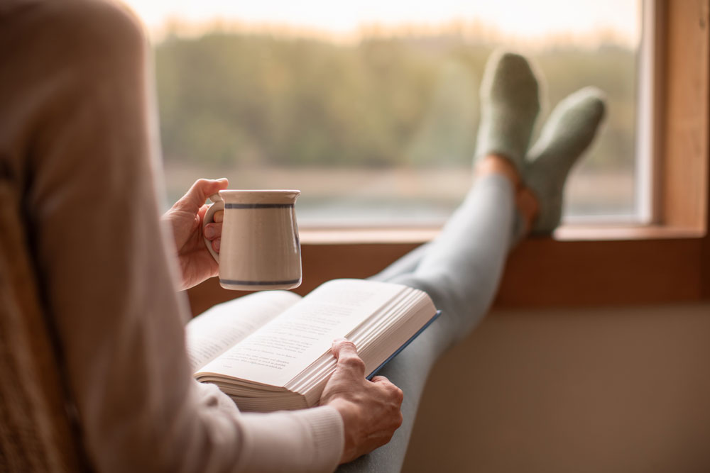 woman enjoying a book and a cup of coffee with her feet up by the window