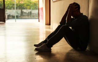 Man sitting on the floor in a dim hallway with his back against the wall and head in his hands.