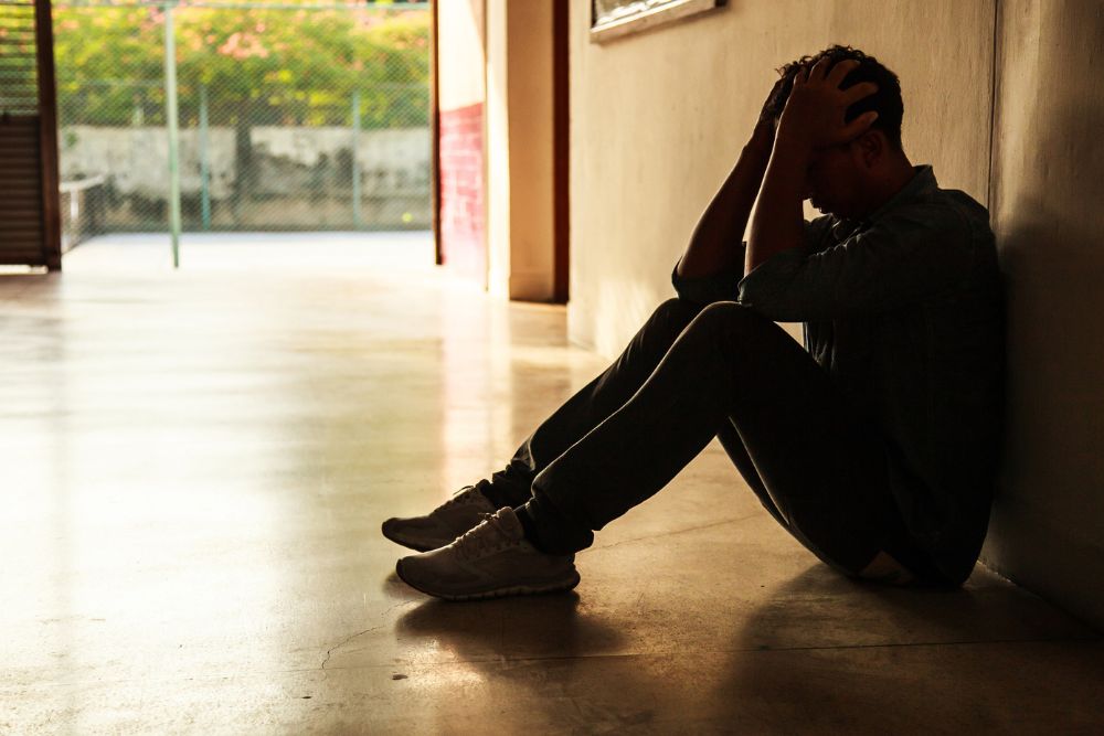Man sitting on the floor in a dim hallway with his back against the wall and head in his hands.