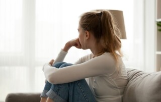 Young woman sitting on a couch with knees drawn up, looking out the window with a thoughtful or concerned expression.