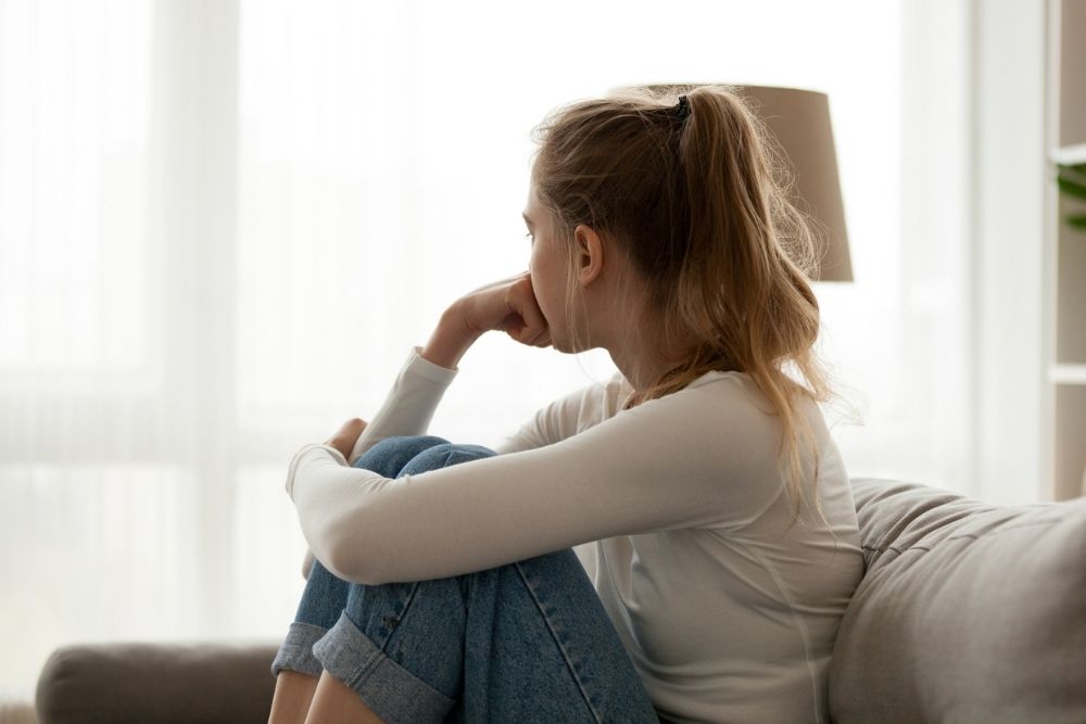 Young woman sitting on a couch with knees drawn up, looking out the window with a thoughtful or concerned expression.