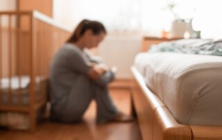 Blurred image of a woman sitting on the floor next to a bed and crib, hugging her knees