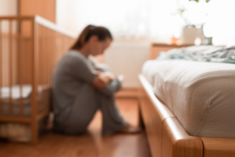 Blurred image of a woman sitting on the floor next to a bed and crib, hugging her knees