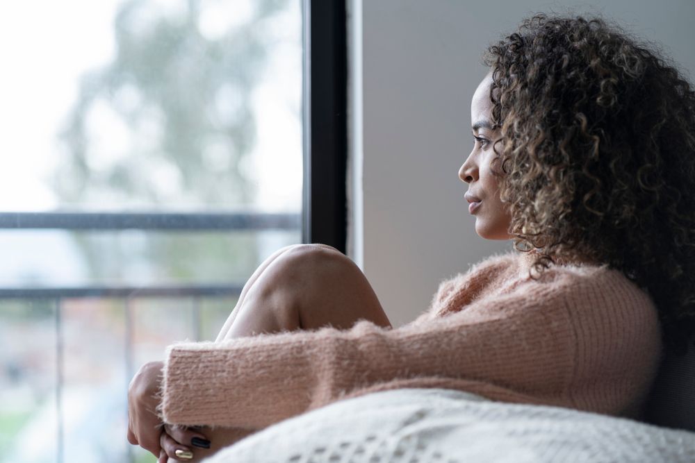Woman sitting indoors with her knees pulled to her chest, looking thoughtfully out a window, wearing a soft pink sweater.