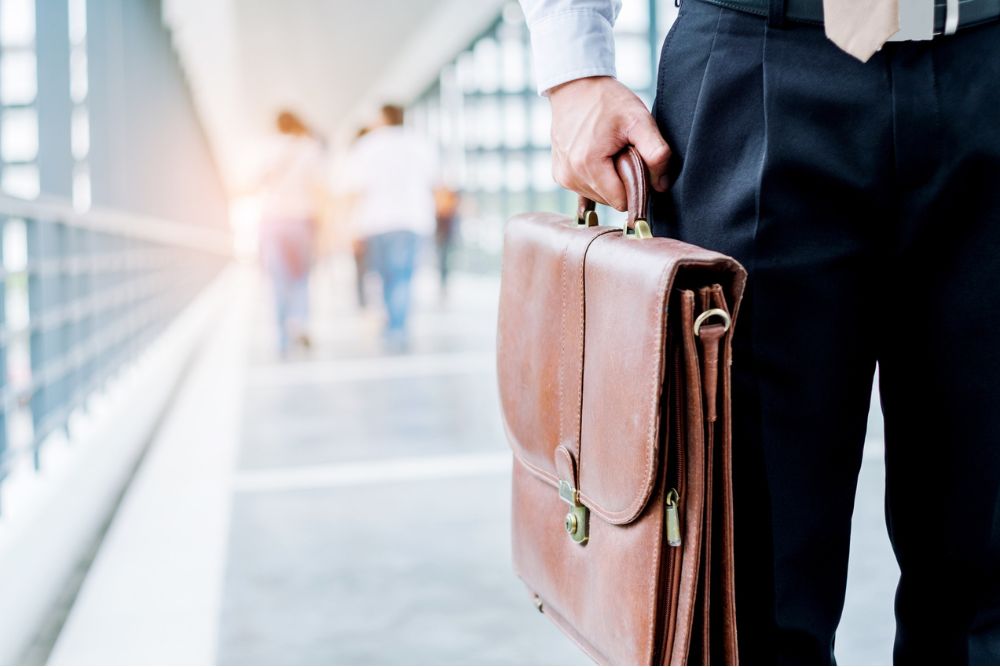 Businessperson holding a leather briefcase while walking in a modern office corridor, symbolizing professionalism, employment, or workplace issues.