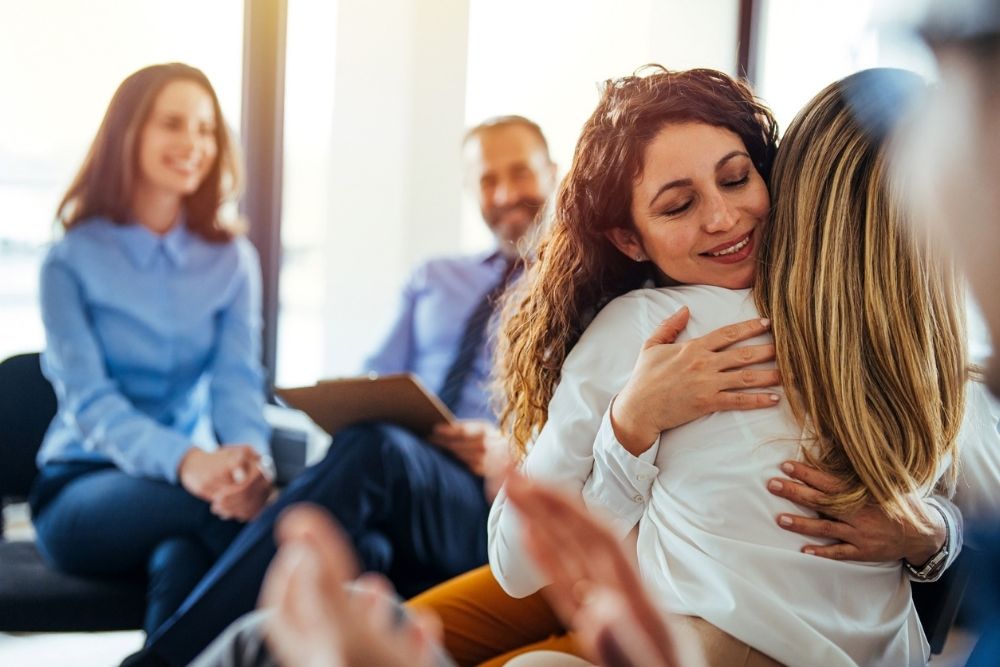 Two women hug warmly in a group setting, smiling with relief and support. Other people sit nearby, watching and clapping. The atmosphere feels positive and encouraging.