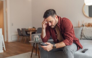 A man sitting on a couch looking at his phone with a distressed expression, holding his forehead, suggesting worry, anxiety, or emotional struggle.