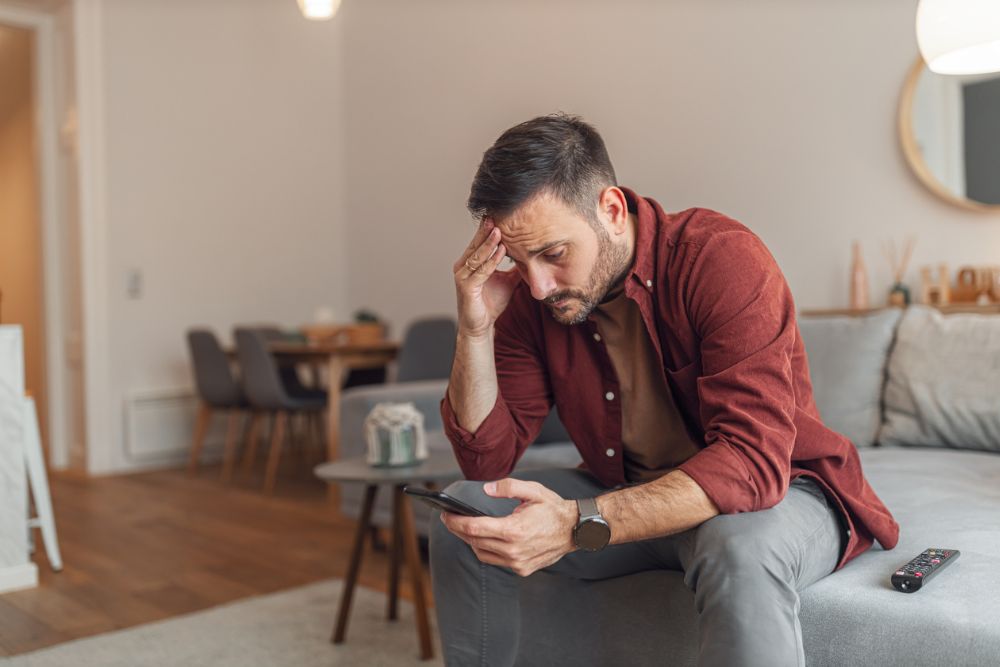 A man sitting on a couch looking at his phone with a distressed expression, holding his forehead, suggesting worry, anxiety, or emotional struggle.