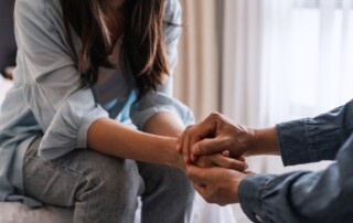 Close-up of two people sitting indoors, one offering comfort by holding the other's hands. The scene conveys emotional support, compassion, and a caring interaction, possibly in a therapeutic or supportive setting.