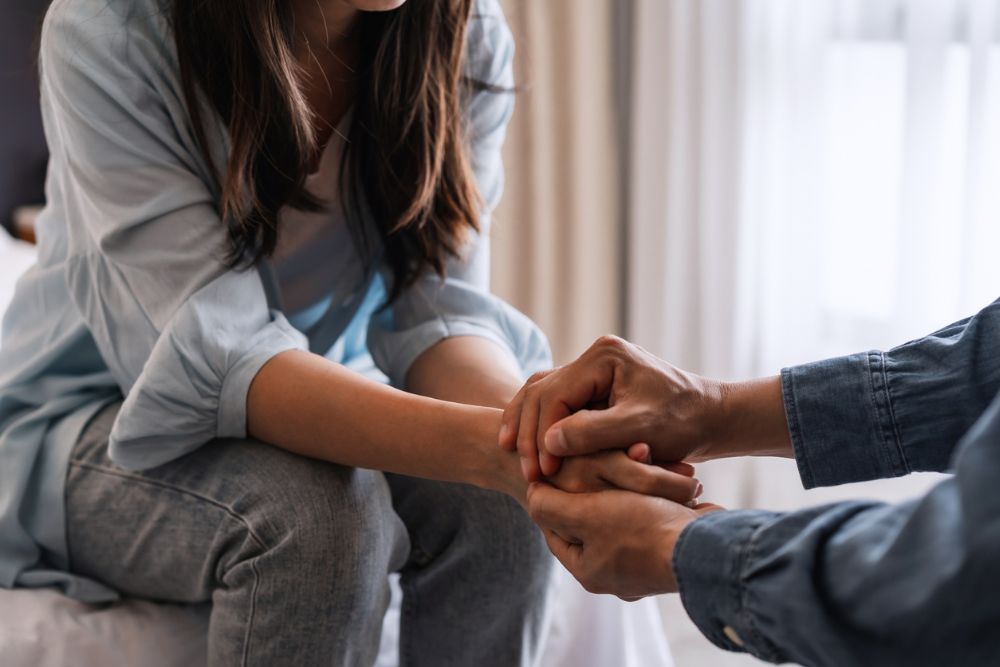 Close-up of two people sitting indoors, one offering comfort by holding the other's hands. The scene conveys emotional support, compassion, and a caring interaction, possibly in a therapeutic or supportive setting.