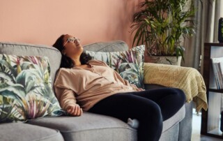 Woman reclining on a couch with her head tilted back and eyes closed, appearing emotionally or physically drained in a cozy living room setting