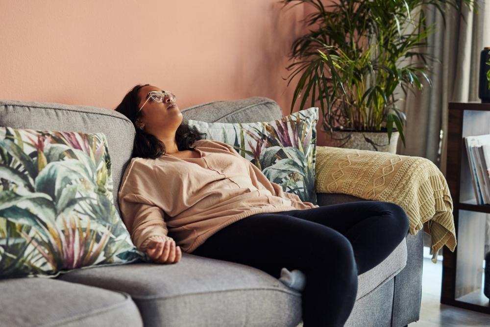 Woman reclining on a couch with her head tilted back and eyes closed, appearing emotionally or physically drained in a cozy living room setting