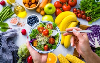 Close-up of a person holding a bowl of fresh salad with cherry tomatoes, leafy greens, and sprouts, surrounded by colorful fruits and vegetables on a kitchen counter.