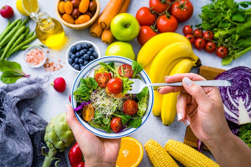 Close-up of a person holding a bowl of fresh salad with cherry tomatoes, leafy greens, and sprouts, surrounded by colorful fruits and vegetables on a kitchen counter.