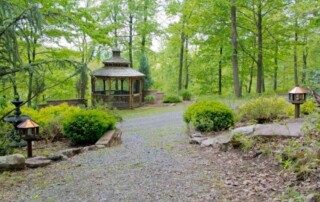 A wooden gazebo surrounded by trees and greenery along a gravel path in a peaceful outdoor setting.