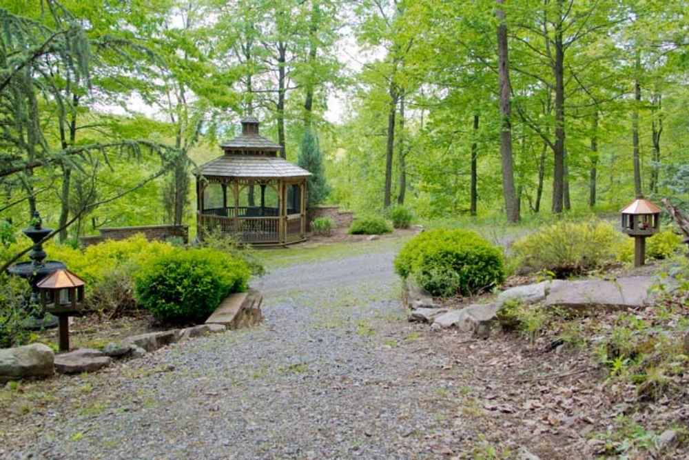 A wooden gazebo surrounded by trees and greenery along a gravel path in a peaceful outdoor setting.