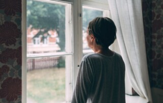 A woman with short hair stands indoors, looking out a window with sheer curtains, appearing thoughtful as she gazes toward houses and trees outside.