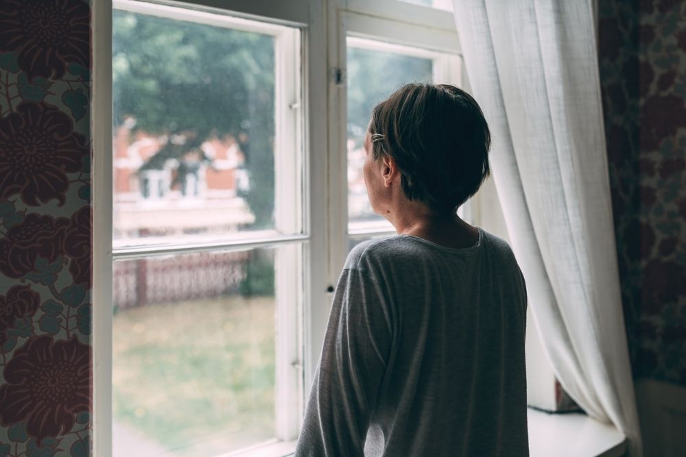A woman with short hair stands indoors, looking out a window with sheer curtains, appearing thoughtful as she gazes toward houses and trees outside.