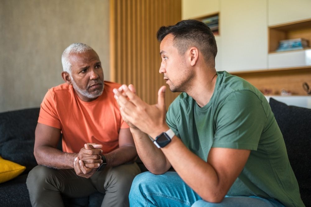 Two men sit on a couch engaged in a serious conversation, with one speaking and gesturing while the other listens attentively.