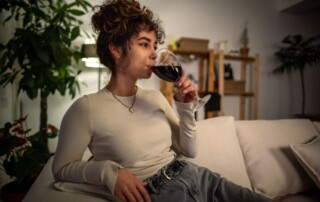 Young woman relaxing on a couch at night while drinking a glass of red wine, symbolizing using alcohol to unwind after a long day.