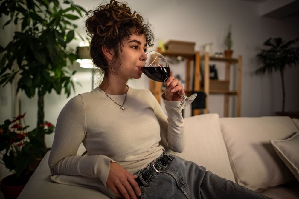 Young woman relaxing on a couch at night while drinking a glass of red wine, symbolizing using alcohol to unwind after a long day.