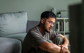 A man sits on the floor indoors with his arms wrapped around his knees, looking thoughtful and concerned.