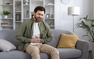 Man sitting on a couch holding his stomach in discomfort, appearing to experience abdominal pain or digestive issues.
