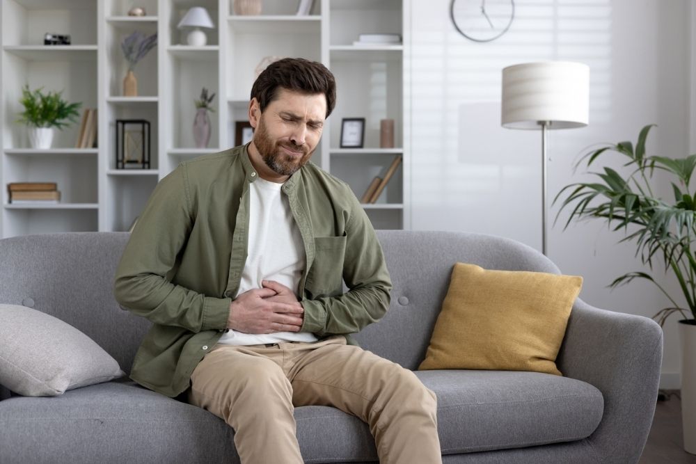 Man sitting on a couch holding his stomach in discomfort, appearing to experience abdominal pain or digestive issues.