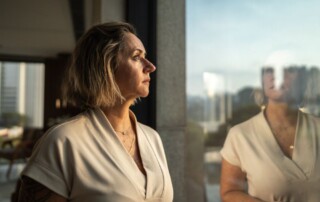 Middle-aged woman standing by a window, looking outside thoughtfully with her reflection visible in the glass.