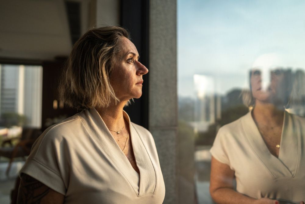 Middle-aged woman standing by a window, looking outside thoughtfully with her reflection visible in the glass.