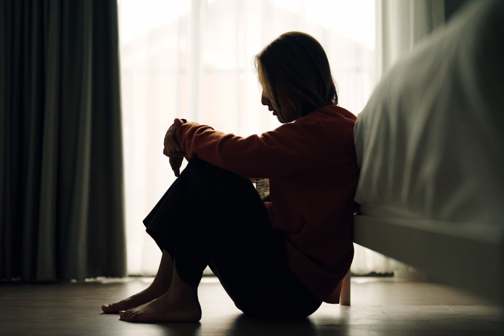 A woman sits on the floor beside her bed in a dimly lit room, her head bowed in sadness, representing emotional struggle and vulnerability.