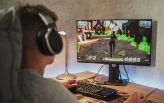 A person wearing headphones sits at a desk playing a video game on a large monitor, focused on a colorful open-world scene.