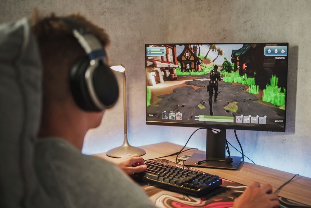 A person wearing headphones sits at a desk playing a video game on a large monitor, focused on a colorful open-world scene.