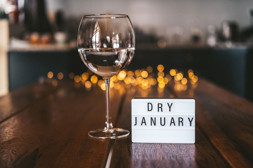 a wine glass sits on a table. A white sign with black border reads dry january in black letters.