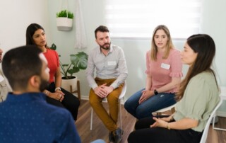 St Joseph - What Is Integrated Care in Recovery. A group of young individuals sit in a circle in a well lit room. Each has a name tag and is listening to a woman in a green shirt speak.