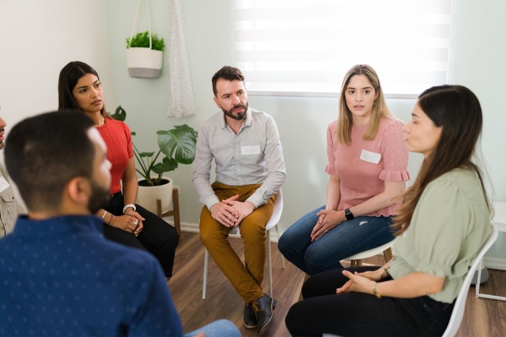 St Joseph - What Is Integrated Care in Recovery. A group of young individuals sit in a circle in a well lit room. Each has a name tag and is listening to a woman in a green shirt speak.